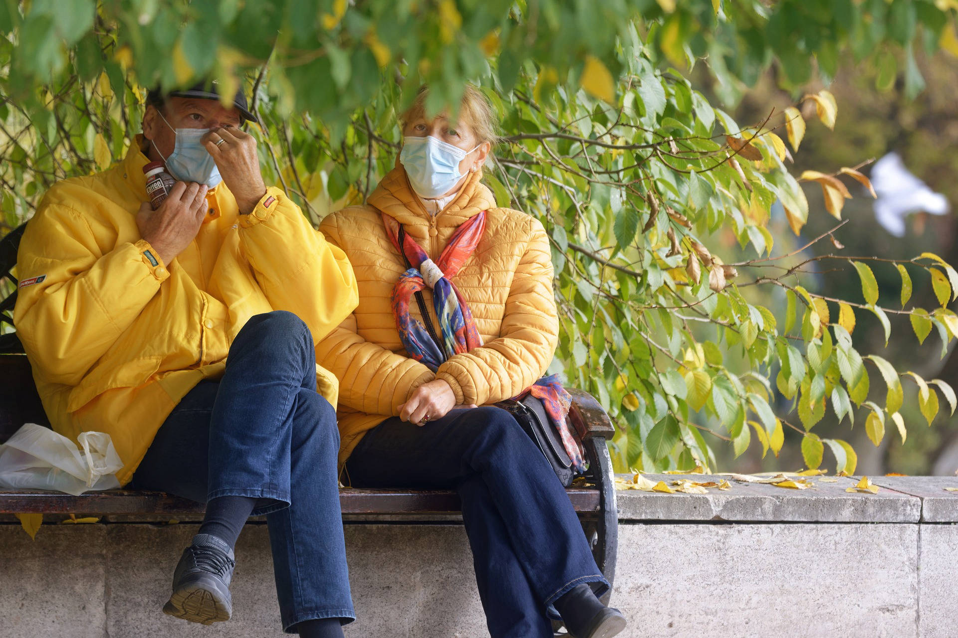 Two people sitting on a bench wearing yellow coats and blue face masks.